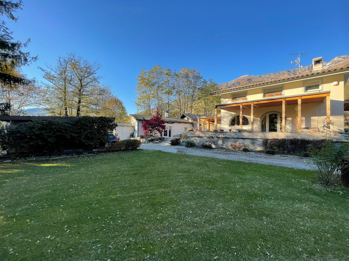 A spacious green lawn surrounded by trees is visible in the foreground. The villa stands to the right, featuring a covered entrance and large windows. In the background, the clear blue sky complements the serene landscape, creating a peaceful outdoor setting.