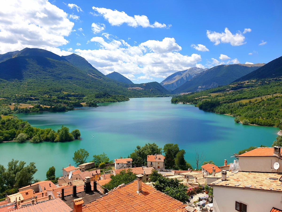 A panoramic view of a tranquil lake surrounded by green hills and mountains, reflecting vivid blue and turquoise hues. The foreground reveals terracotta rooftops of a charming village, adding to the serene landscape under a partly cloudy sky.