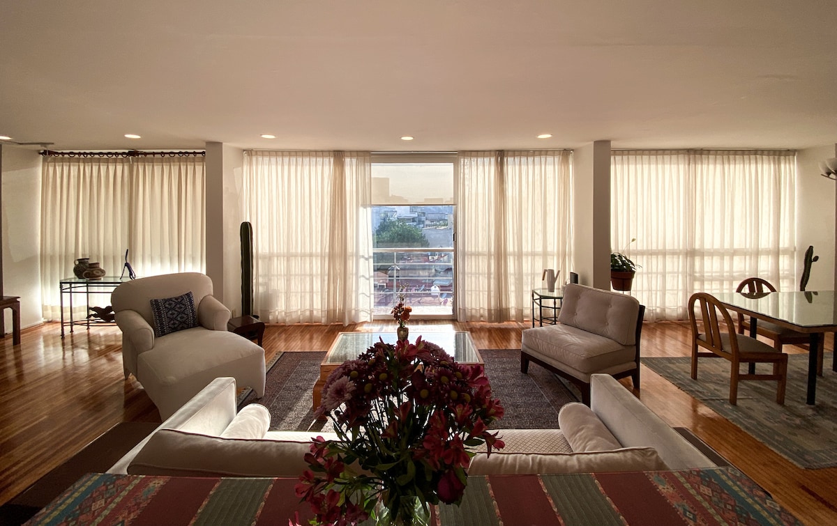 A bright living room features large windows with sheer curtains, allowing natural light to fill the space. Comfortable seating includes a sofa and armchair, complemented by a coffee table adorned with flowers. A dining table is positioned nearby, with warm wooden flooring adding to the ambiance.