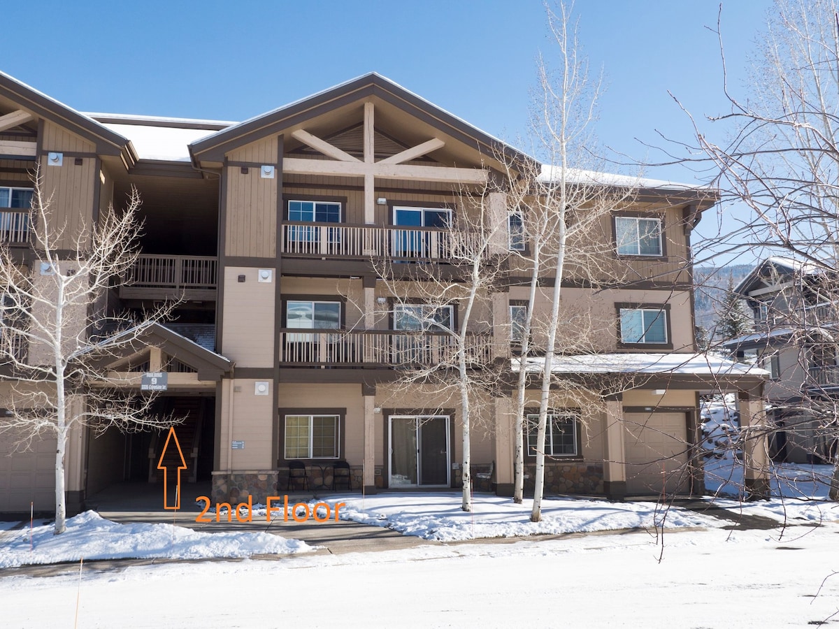 The exterior of a three-story building is shown, featuring a combination of light and dark tan siding. The second floor is highlighted with an arrow, indicating the unit's entrance. Snow covers the ground, and surrounding trees are bare, signaling winter.