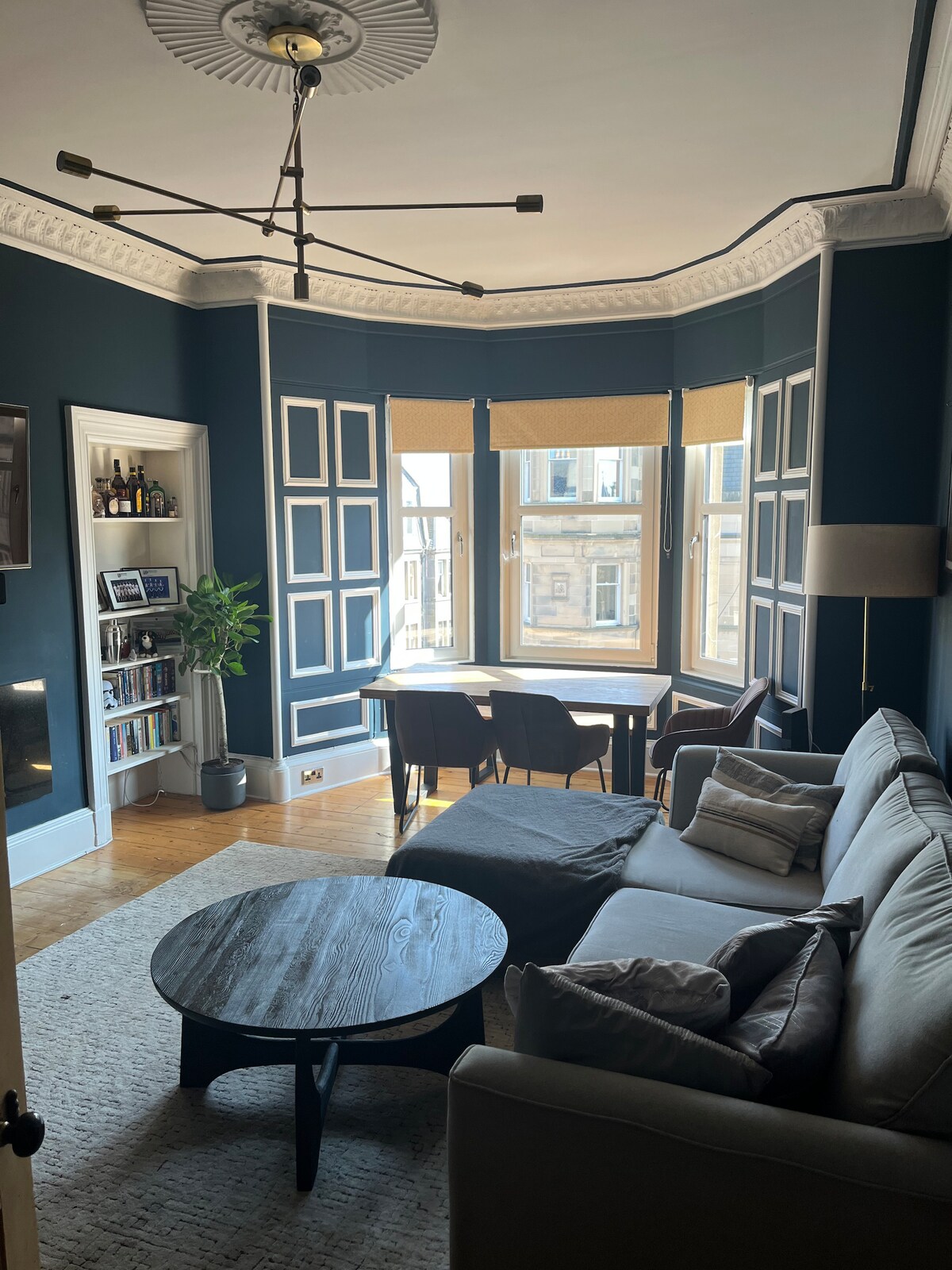 A spacious living room features a large gray sectional sofa with decorative pillows, positioned near a round wooden coffee table. Natural light filters through tall windows adorned with beige shades, illuminating the dark blue walls and creating a cozy atmosphere. A dining area is visible, with a wooden table surrounded by chairs.