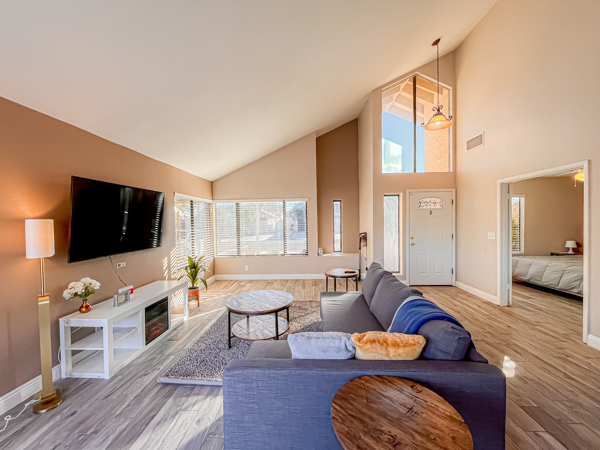 A spacious living room features large windows allowing natural light to fill the area. A gray sectional sofa is arranged around a circular coffee table on a soft rug. A flat-screen TV is mounted on the wall opposite the seating, enhancing the cozy atmosphere.