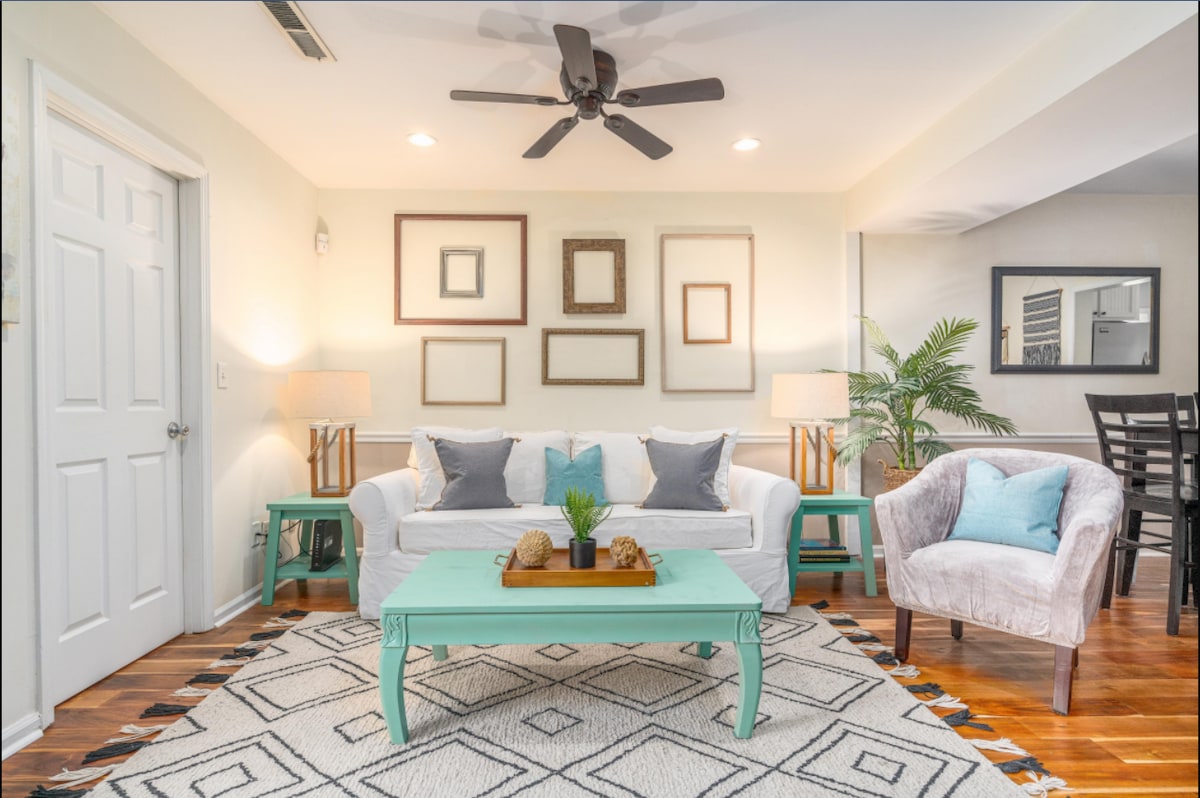 A light-filled living room features a white sofa adorned with blue and gray cushions, complemented by a cozy armchair and a turquoise coffee table. Framed art is displayed on the wall above the sofa, while a ceiling fan circulates air in the welcoming space.
