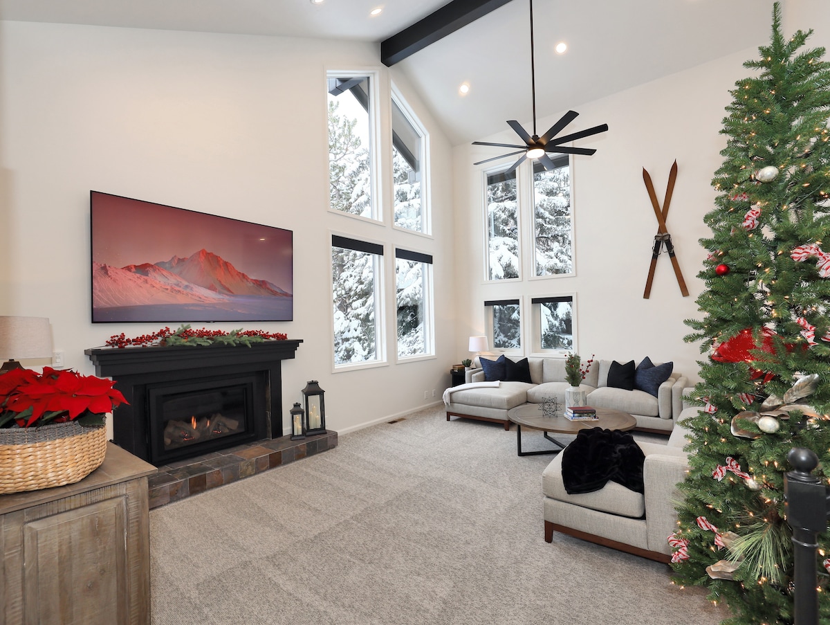 A spacious living area features large windows that allow natural light and showcase snowy outdoor views. A modern fireplace is framed by a comfortable seating arrangement. A decorative Christmas tree is positioned nearby, adding a festive touch to the environment.