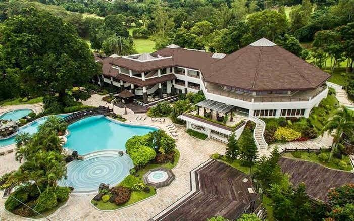 An aerial view showcases a sprawling vacation home surrounded by lush greenery. A large swimming pool is visible, complemented by landscaped gardens and outdoor seating areas. The architectural design features multiple levels with a distinctive roof and ample outdoor space for relaxation and recreation.