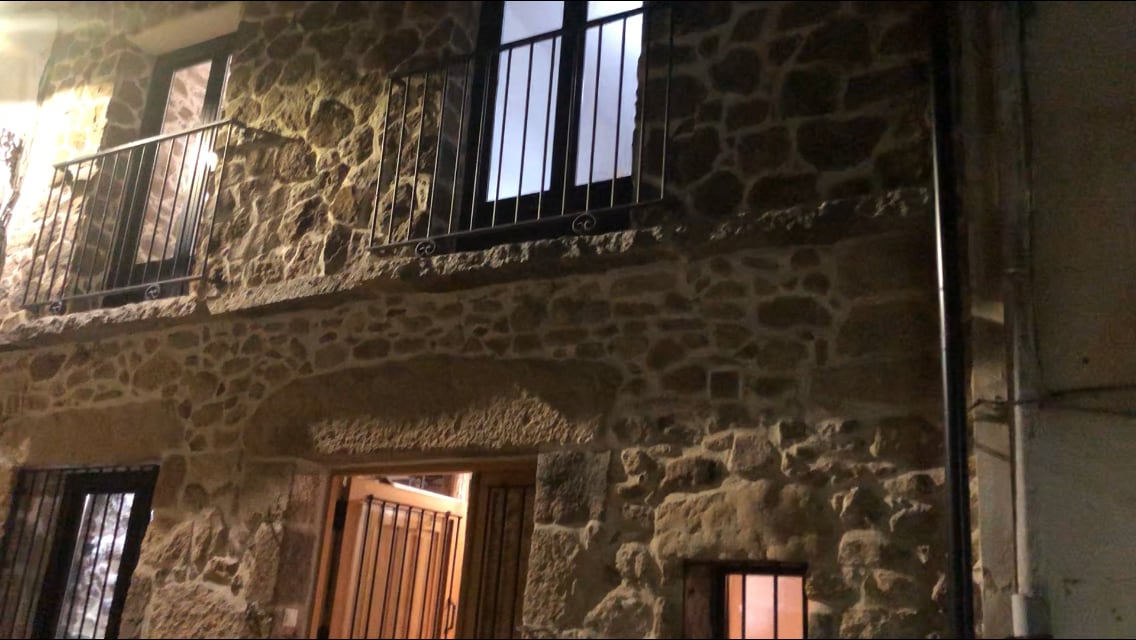 The exterior view showcases a rustic stone facade with two upper windows framed by black railings. A wooden door leads into the building, revealing warm light from within. The texture of the stone adds character to the inviting entrance.