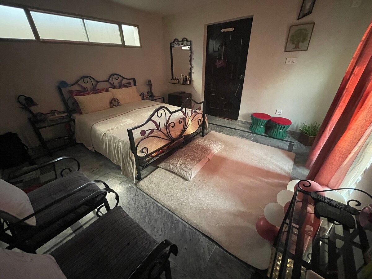 A cozy bedroom features a spacious bed adorned with light bedding. A soft rug is placed on the floor with floor cushions nearby. The room is illuminated by natural light streaming through a large window, while decorative elements and a mirror add to the inviting atmosphere.