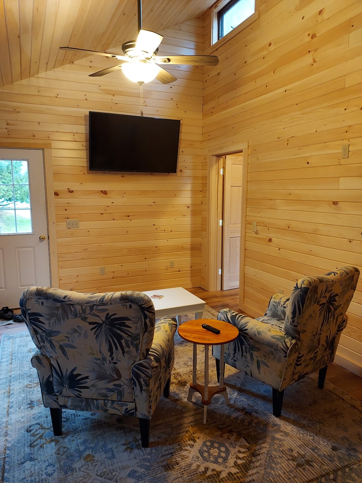 A cozy seating area is revealed, featuring two upholstered armchairs with a tropical leaf pattern. A small side table stands beside them, and a flat-screen television is mounted on the wooden wall. Natural light enters through the nearby door.