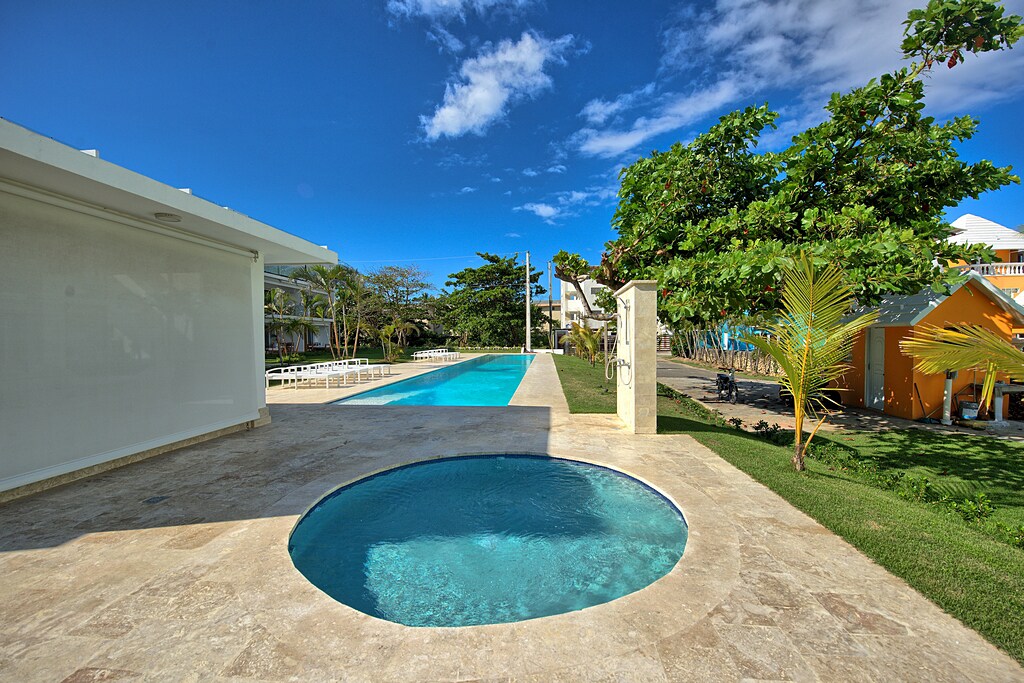 A spacious pool area features a large swimming pool, flanked by lounge chairs under the sun. A circular hot tub is located nearby, with a smooth stone deck surrounding it. Lush greenery and colorful landscaping add a natural touch to the setting.