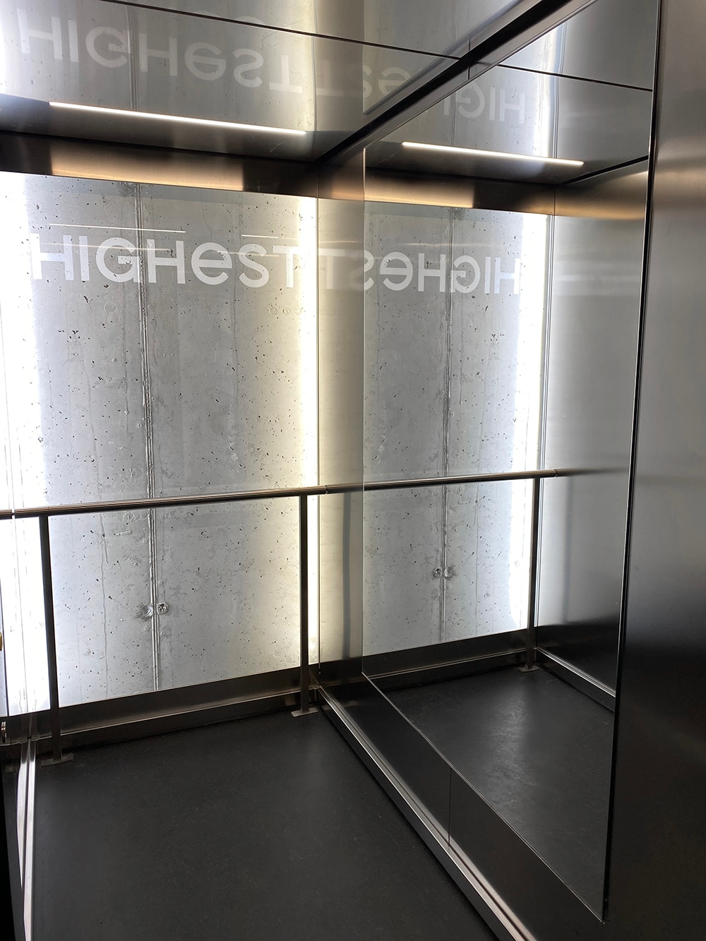 An elevator interior is shown, featuring sleek metal walls and a mirrored back. The words 'HIGHEST' are displayed prominently across the back wall. A handrail runs along one side, providing stability for passengers. The floor is finished in a dark color, contributing to a modern aesthetic.