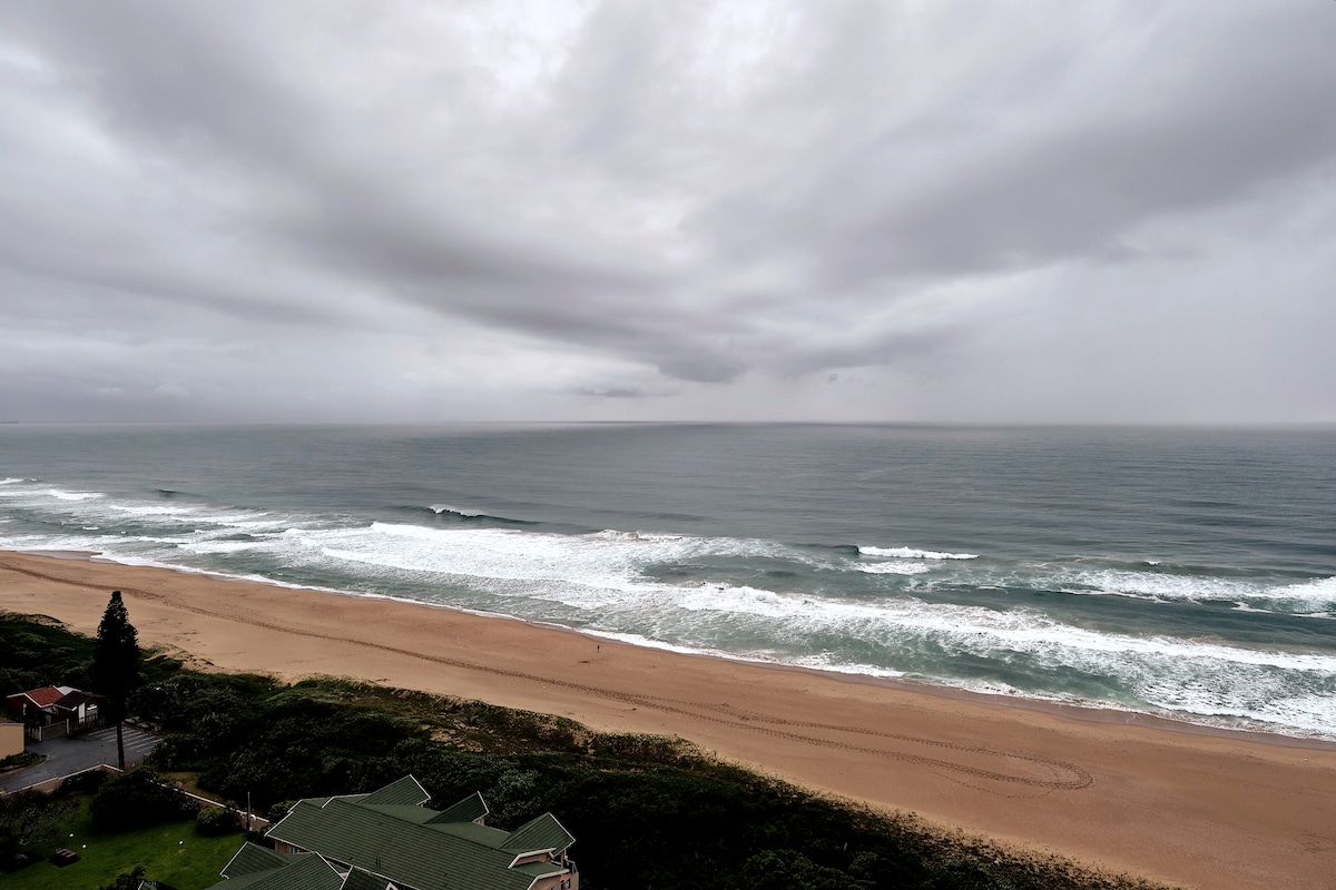 A serene view of a sandy beach leads to gentle ocean waves under a cloudy sky. The shoreline stretches along the coast, with soft waves lapping at the sand. A green-roofed building is visible in the foreground, surrounded by lush vegetation.