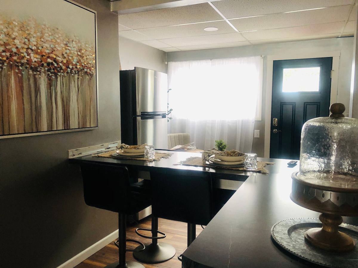 The dining area features a wooden table set for two, accompanied by black bar stools. A modern refrigerator is positioned nearby. Natural light fills the space through a large window covered by sheer white curtains, enhancing the overall ambiance.