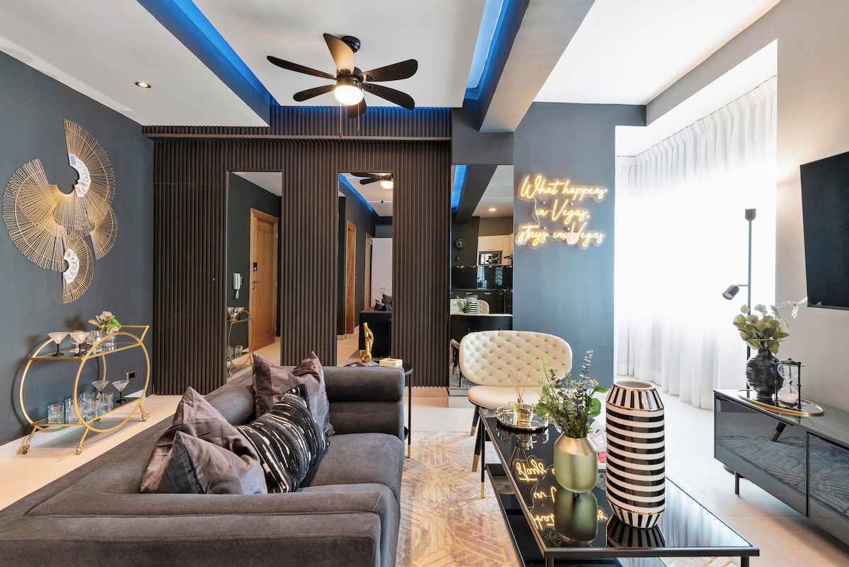 A modern living area features a comfortable sectional sofa adorned with decorative pillows. A stylish coffee table sits in front, complemented by a decorative vase. Soft lighting highlights the sleek wall accents and a neon wall sign, creating an elegant ambiance. Natural light filters through sheer curtains.