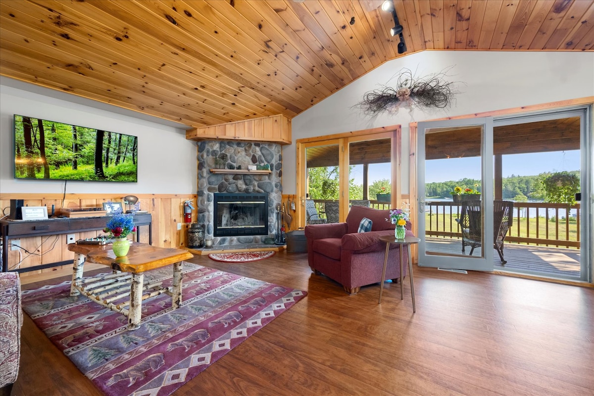 The open living area features wooden ceilings and a stone fireplace, enhancing a cozy feel. A large screen displays a nature scene. Floor-to-ceiling glass doors provide views of the outdoor balcony and Kayuta Lake, allowing natural light to fill the space.