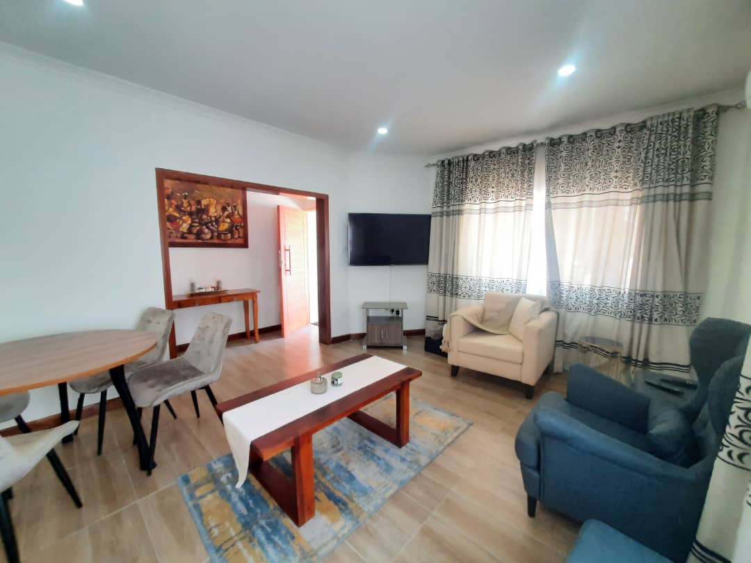 A light-filled living area features a wooden coffee table placed on a decorative rug. A cozy blue armchair and a beige armchair are arranged around the table. A television is mounted on the wall, with large windows framed by patterned curtains.