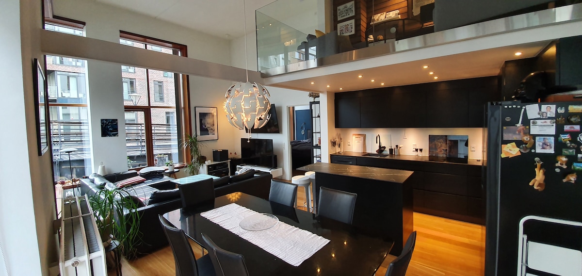 An open-plan living area is depicted with modern furnishings. A sleek black dining table is centered under a distinctive light fixture. The kitchen area is visible, featuring dark cabinetry and ample counter space. A cozy seating arrangement is situated near large windows, allowing natural light to fill the space.
