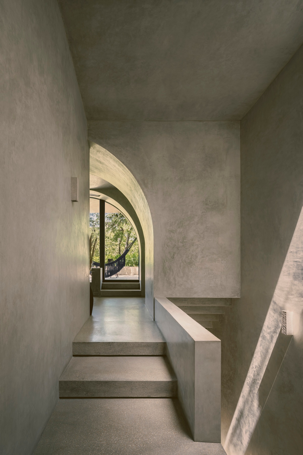 A minimalist stairway leads through an arched passageway, with natural light illuminating the textured walls. A view of the outdoor greenery is framed by the large window at the end, creating a serene connection to the environment.