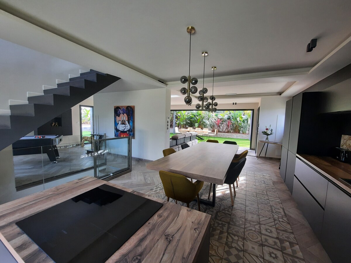A modern kitchen and dining area features a central wooden table surrounded by colorful chairs. A sleek kitchen design includes an island with a stovetop. Large glass doors provide views of the landscaped garden and bring in natural light.