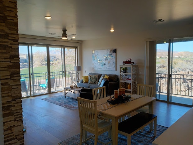 An open concept living area features large windows that provide a view of the golf course. A comfortable sofa is positioned near the glass doors, with a dining area that includes a table and chairs. Natural light enhances the wood flooring and wall decor.