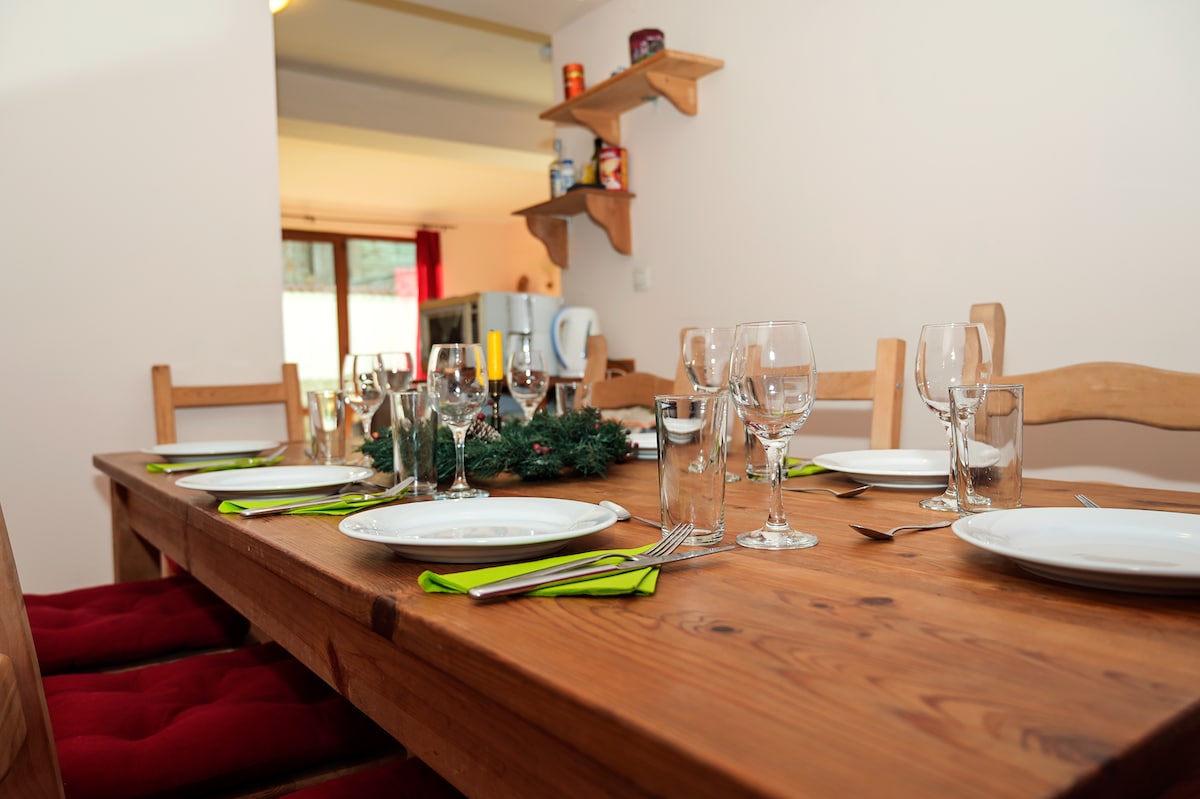 A large wooden dining table is set with plates, glasses, and utensils, creating an inviting dining space. Red cushions adorn the chairs for comfort. The background features shelves with kitchen supplies, while soft light comes from a nearby window, enhancing the ambiance.