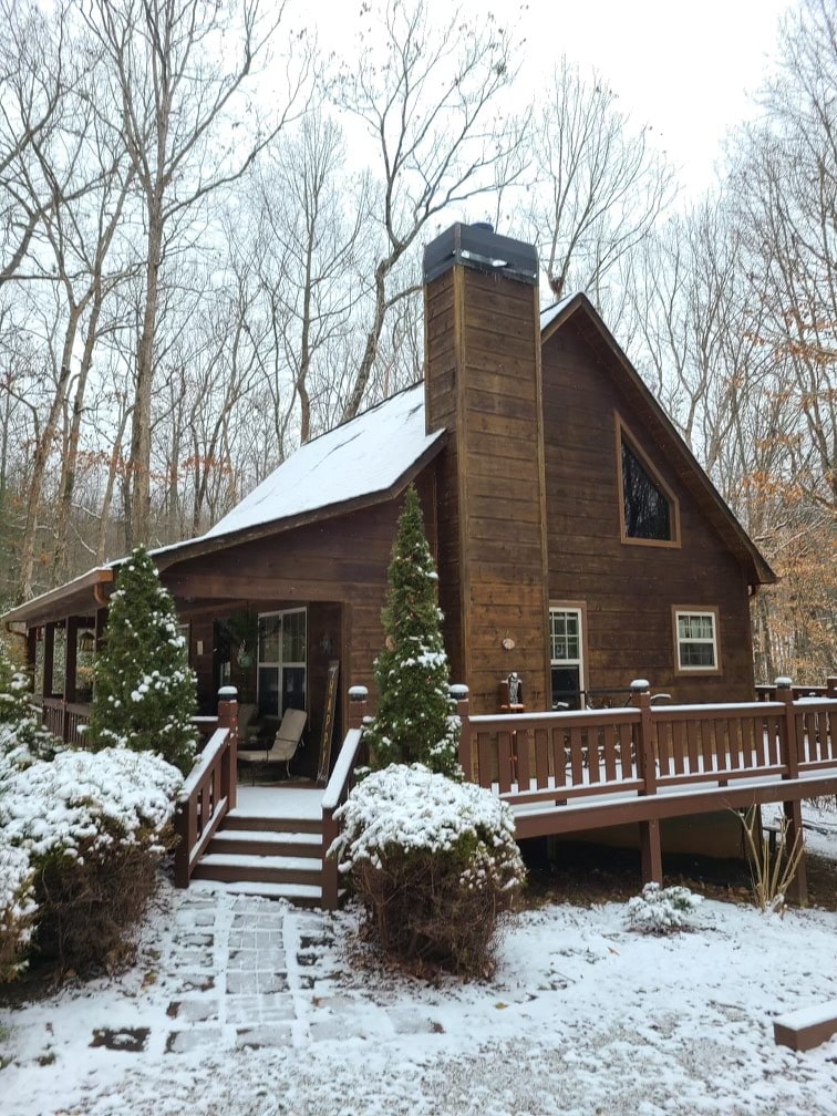 A rustic wooden cabin is surrounded by snow-covered trees, featuring a prominent stone chimney. A spacious wrap-around porch is adorned with neatly placed patio furniture. Lush green shrubs frame the entrance, while a pathway leads to the front door.