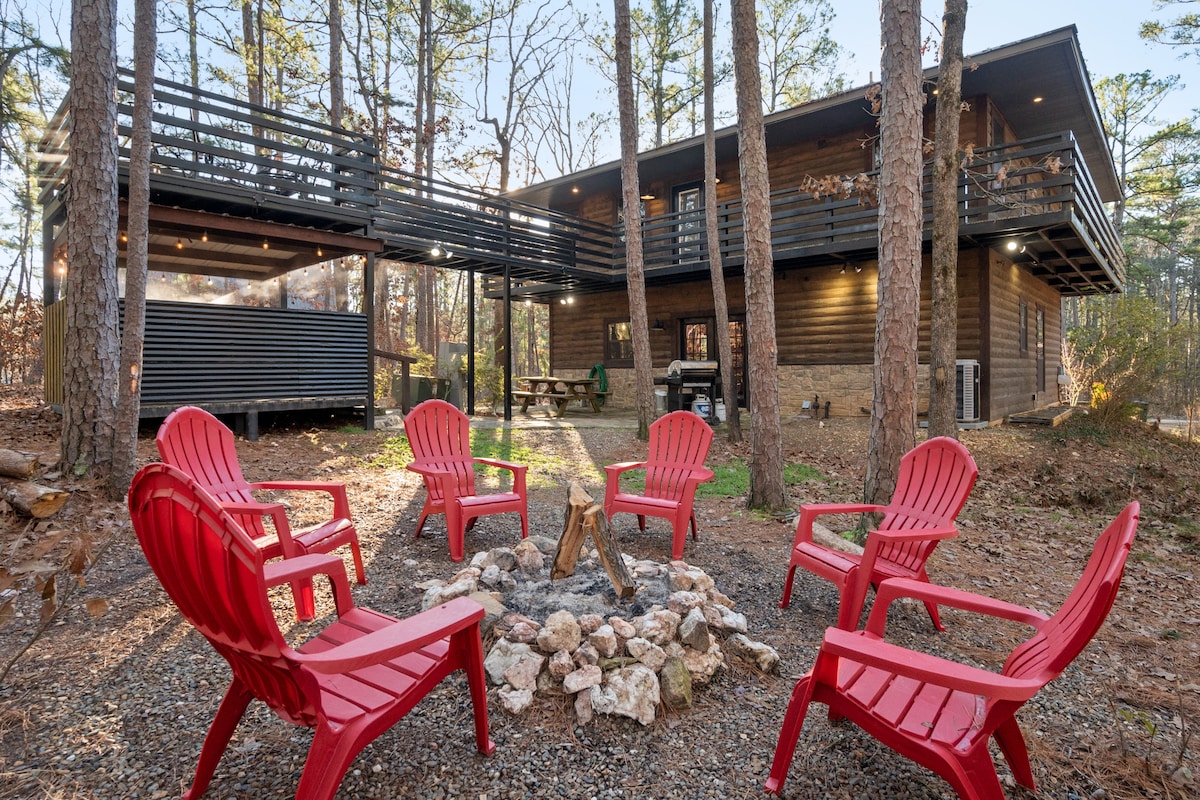 An outdoor fire pit area features several red Adirondack chairs arranged in a circle around a stone fire ring. The cabin stands in the background, partially surrounded by trees, with a wraparound balcony visible above.