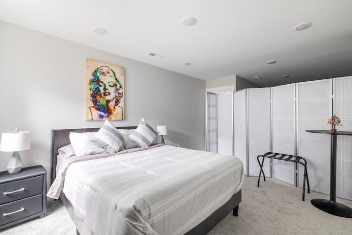 غرفة نوم في الطابق السفلي بسرير مريح بحجم كوين. يتم تقسيم هذه الغرفة بواسطة فاصل جداري. على الرغم من أنه لا يحتوي على جدار تقليدي ، إلا أنه لا يزال يبدو كمنطقة خاصة.