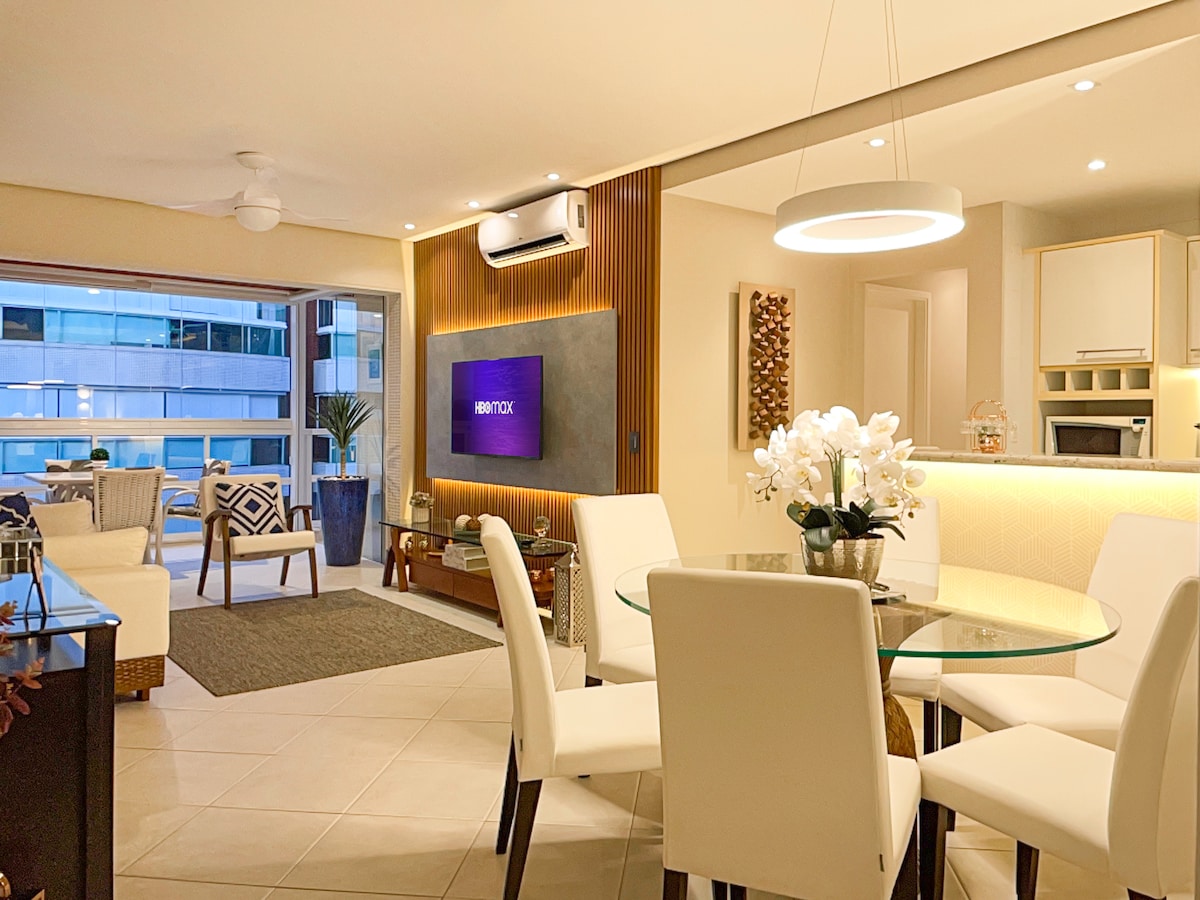 An inviting dining area features a circular glass table surrounded by six white chairs. Soft lighting illuminates the space, which includes a television on the wall. A comfortable seating area with a plush couch and decorative accents is visible in the background, alongside large windows providing natural light.