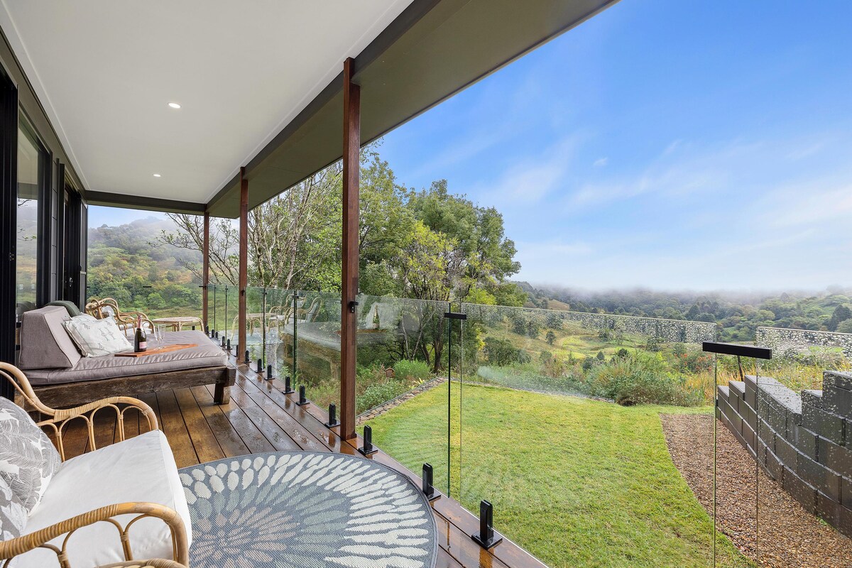 An inviting undercover timber deck features a large daybed and comfortable seating, surrounded by glass balustrades. Lush greenery and expansive valley views extend into the horizon, creating a serene outdoor space perfect for relaxation.
