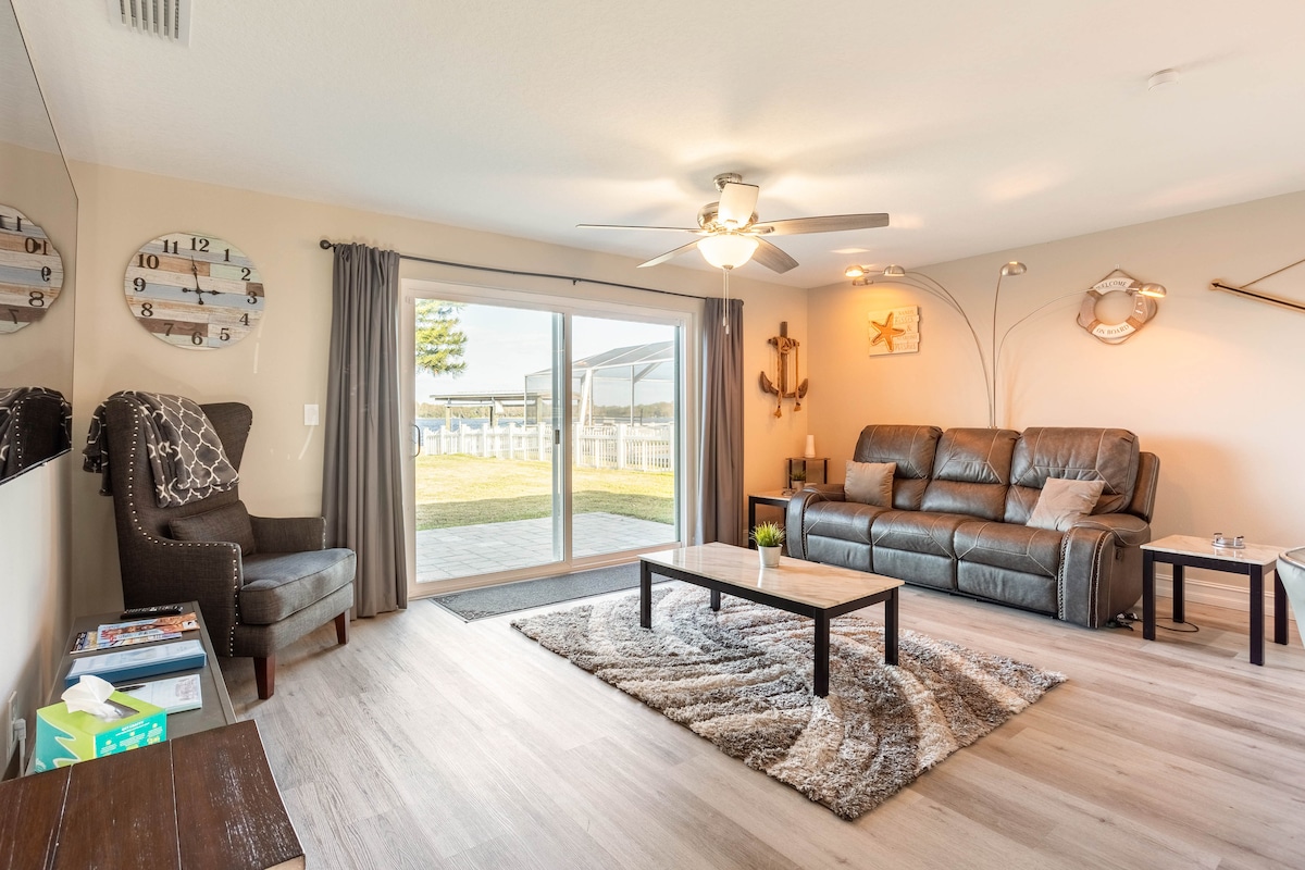 A cozy living area features a comfortable leather couch and an armchair positioned near a large sliding glass door. A coffee table rests on a soft area rug, while natural light fills the space, highlighting the light-colored walls and decorative nautical accents.