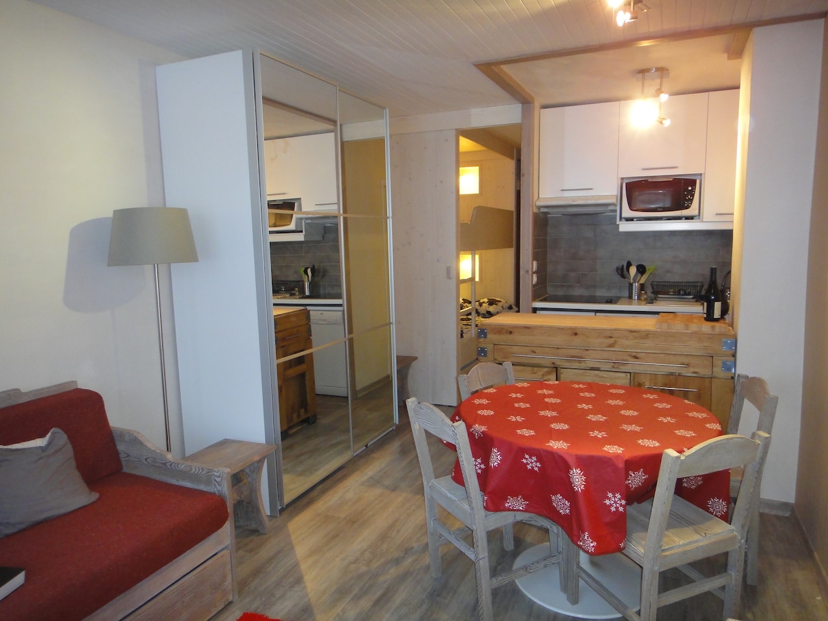 A cozy studio space features a red sofa and a circular dining table with a festive tablecloth. The open kitchen area showcases modern appliances. A mirrored wardrobe reflects the room's light, while subtle lighting creates a serene ambiance. The wood accents add warmth to the decor.