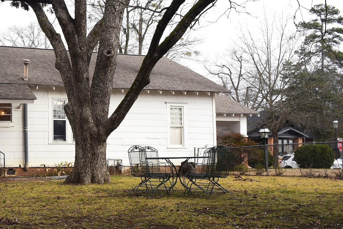 A shaded outdoor space is created under a large tree, featuring a small table with two metal chairs. The yard is primarily grass, surrounded by a fenced perimeter. The white siding of the bungalow is visible in the background, along with hints of nearby homes.