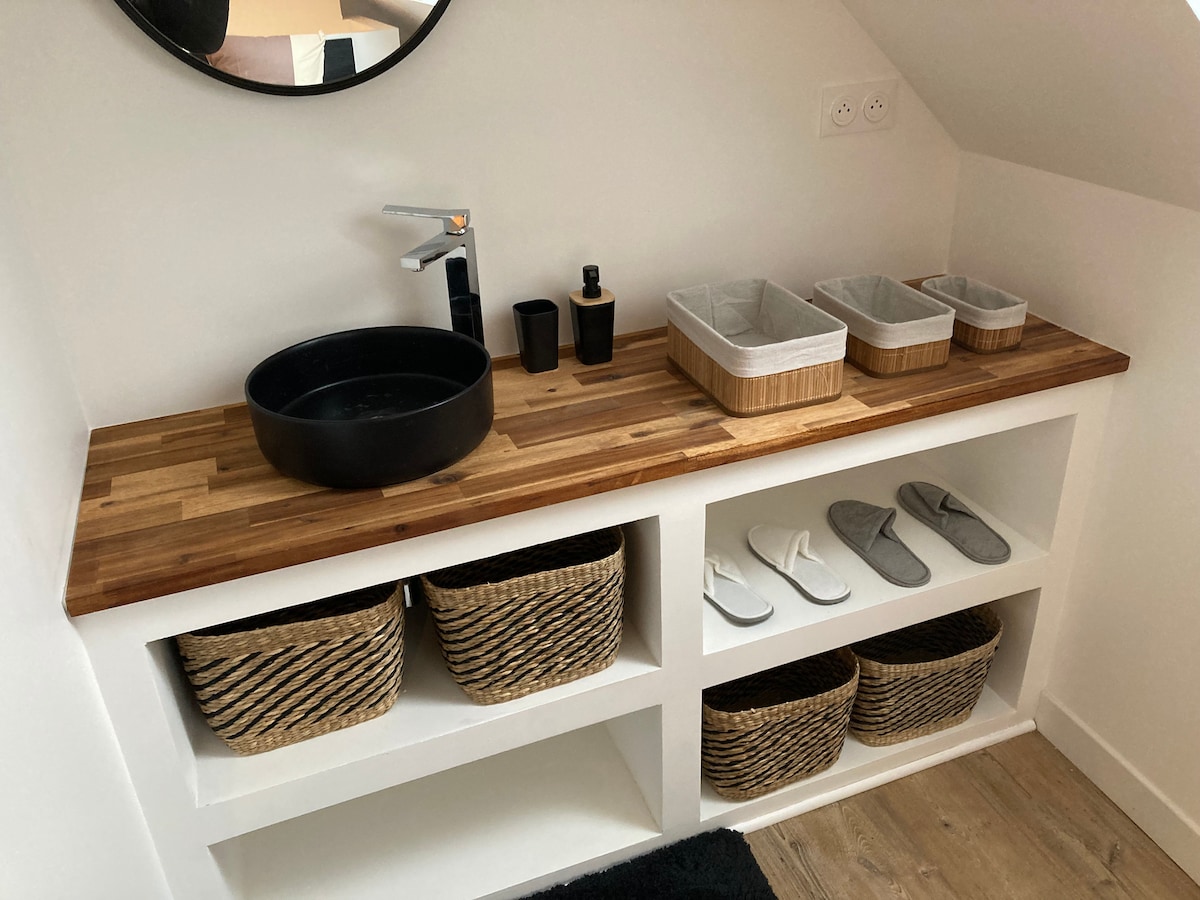 A wooden countertop is presented above a clean white cabinet, featuring a round mirror and a modern black faucet. Neatly arranged baskets hold toiletries and slippers, while two open shelves below offer additional storage with woven baskets.
