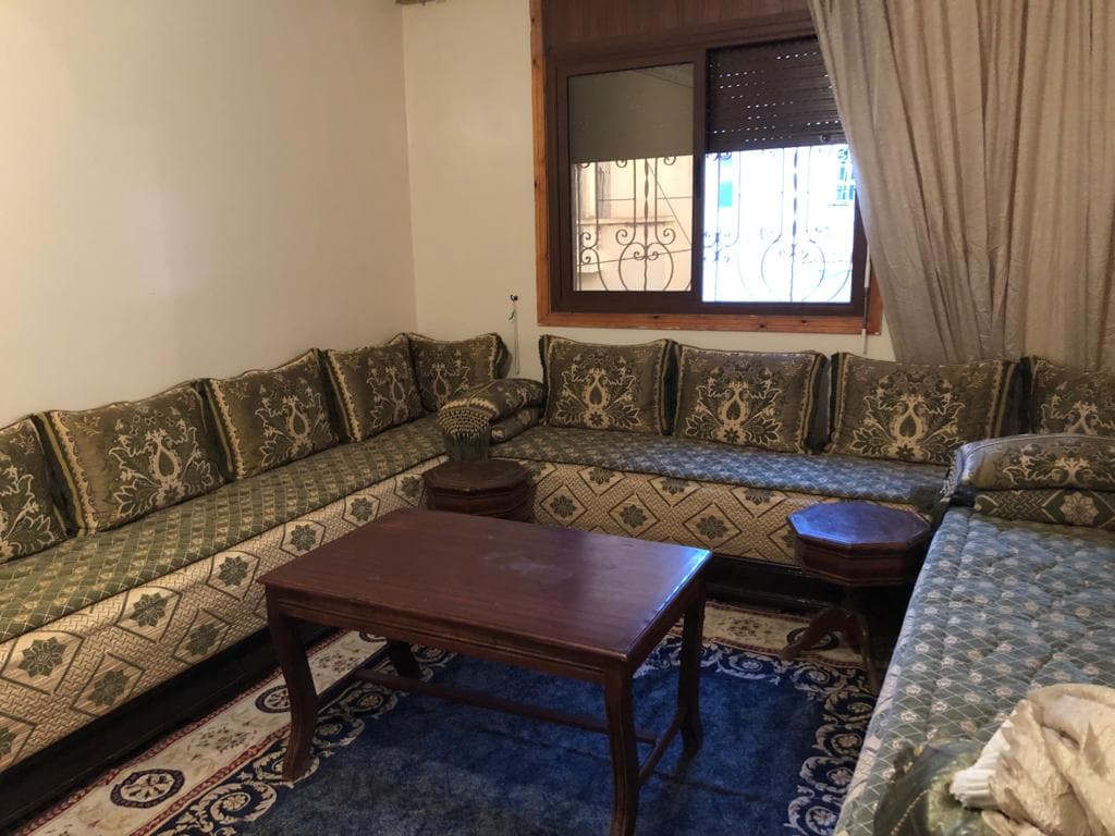 A comfortable seating area showcases intricately patterned cushions arranged along a low table. Natural light enters through a window, illuminating the room's neutral tones and highlighting the decorative rug beneath. A partially draped curtain adds a touch of texture to the space.