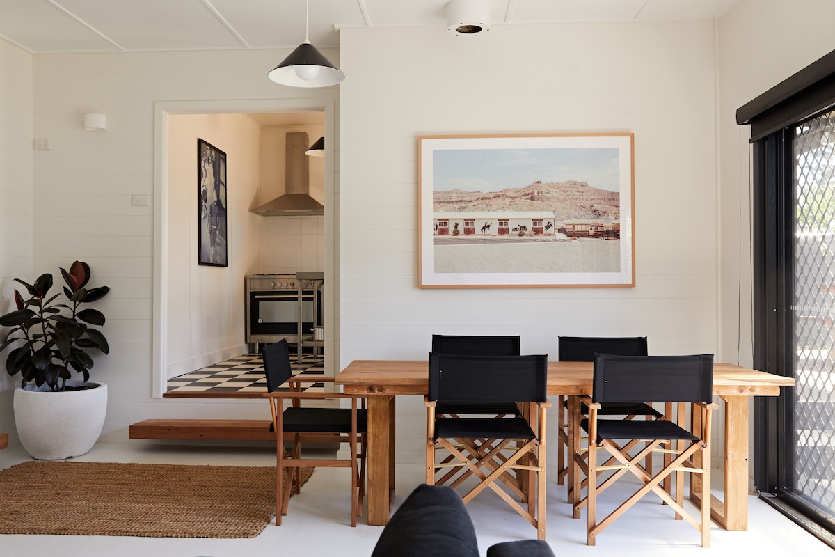 A modern dining area features a wooden table surrounded by black folding chairs. A large artwork hangs on the wall, depicting a scenic landscape, while a doorway leads to a well-equipped kitchen with checkerboard flooring. Indoor plants add a touch of greenery.