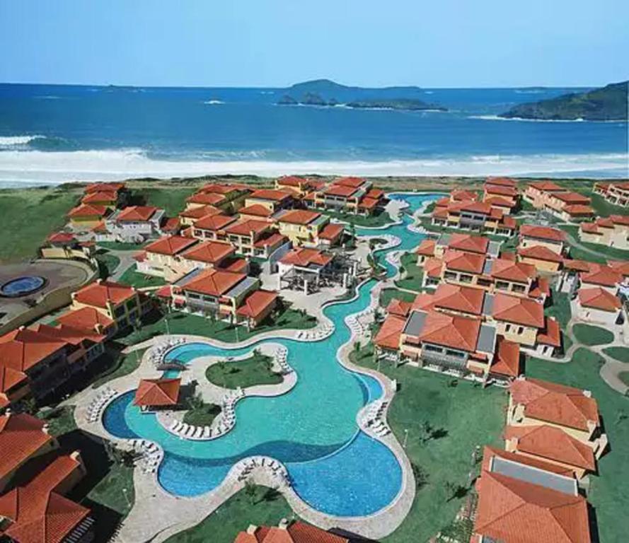 The aerial view captures a sprawling resort with red-roofed villas surrounding a winding pool. The clear water reflects the sunny sky, while the beach and lush greenery are visible in the background, creating a serene coastal atmosphere.