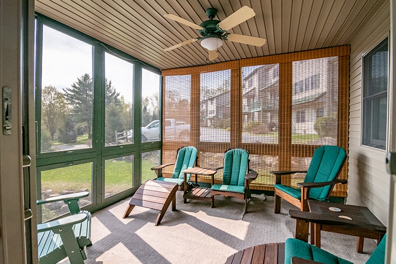 The covered screen porch features teal seating and wooden accent tables, providing a comfortable space for relaxation. Sunlight filters through the screens, illuminating the area and showcasing views of the surrounding greenery.