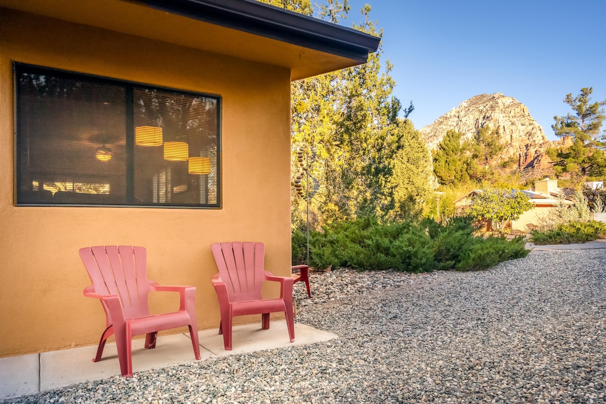 Two red Adirondack chairs are positioned near the home’s entrance, accompanied by a gravel path and surrounded by low shrubs. A mountain peak rises in the background, framed by golden sunlight filtering through foliage.
