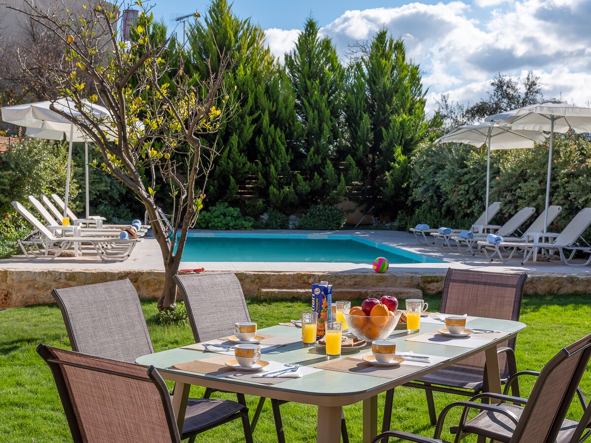 An outdoor dining table is set with cups and orange juice, overlooking a well-maintained garden. Brightly colored fruit sits center stage. In the background, a private pool is surrounded by sun loungers and shaded by umbrellas, amidst lush greenery.