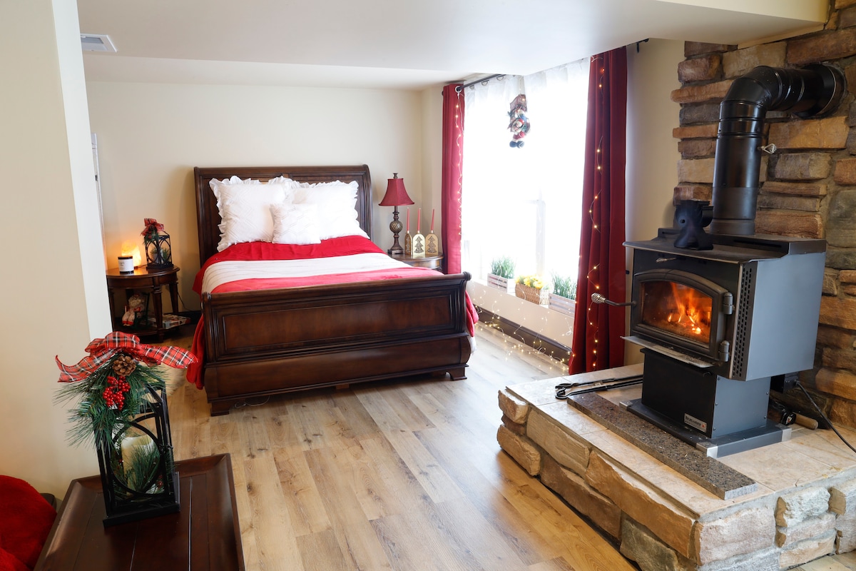 A cozy bedroom features a spacious wooden bed with crisp white linens and a red throw blanket. A wood-burning stove is positioned next to a stone hearth, providing warmth. Warm lighting enhances the ambiance, while a window is adorned with soft curtains and decorative plants.