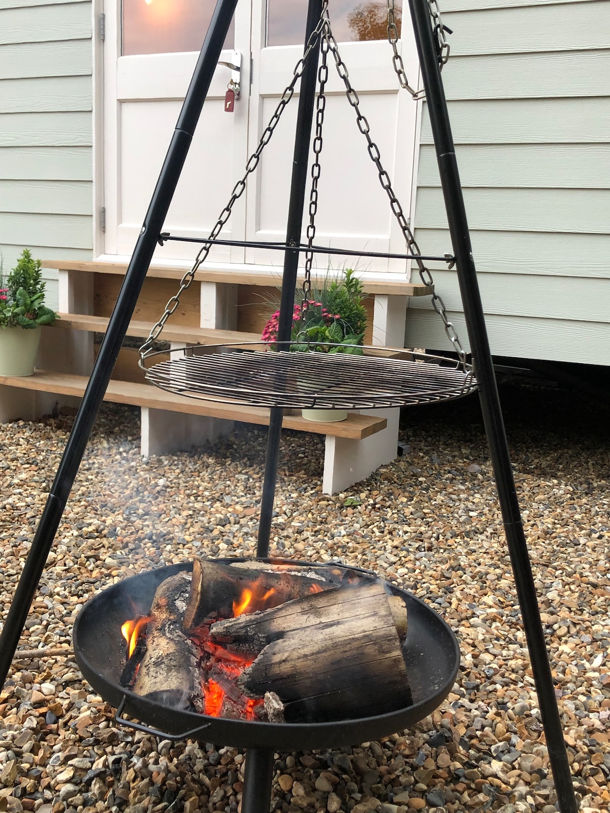 A suspended barbecue grill hangs over a fire pit, where flames are visible consuming logs. The textured gravel ground surrounds the area. In the background, a door leading into the shepherd's hut is partially open, with potted plants positioned nearby.