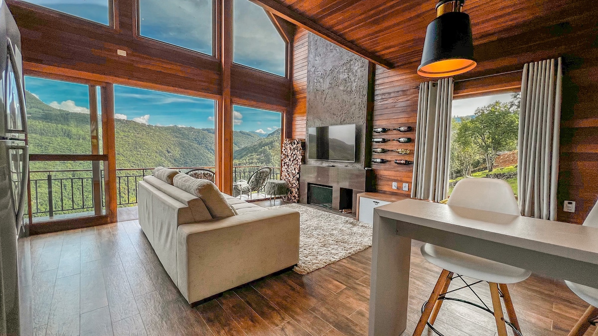 A spacious living area features large windows that provide expansive views of the surrounding hills. A comfortable sofa is positioned near a modern entertainment unit, with a light rug beneath. The dining area includes a simple table and sleek chairs, enhancing the open atmosphere.