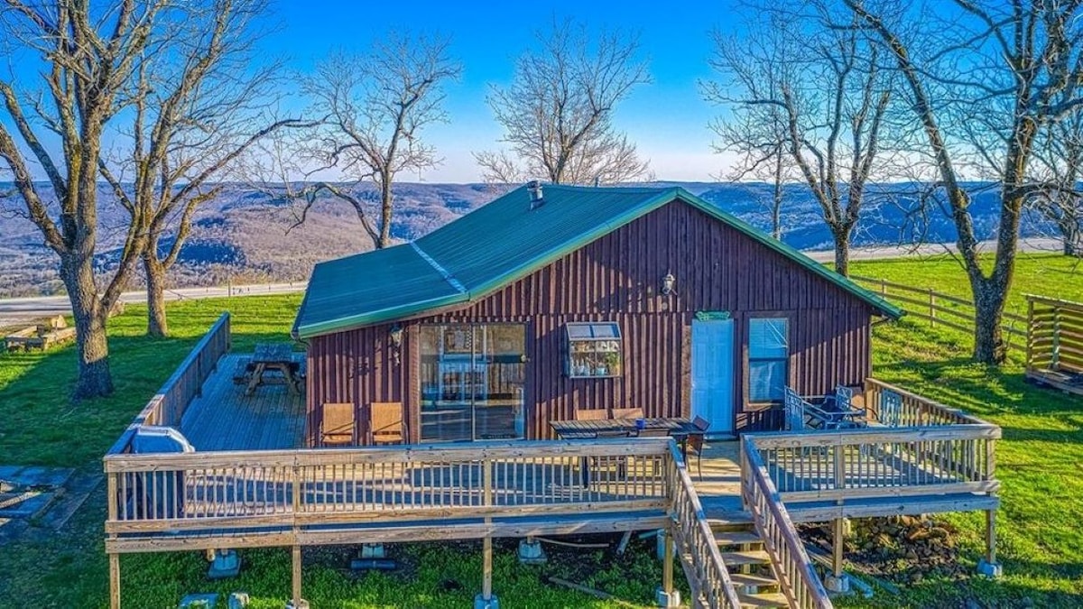 The cabin is shown from an elevated view, featuring a green roof and a spacious wraparound deck with seating. Lush green grass surrounds the structure, and expansive views of rolling hills can be seen in the background beneath a clear blue sky.