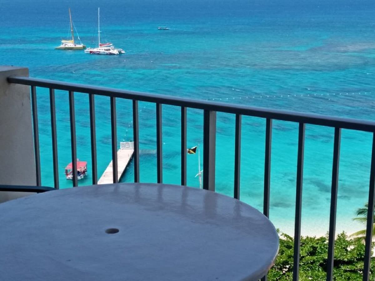 A view from a private balcony showcases the vibrant blue waters of the Caribbean Sea. Boats are seen floating in the distance, while a wooden pier extends toward the water. The railing frames the serene coastal scene, emphasizing the tranquil atmosphere.