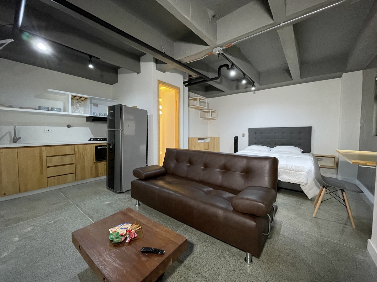 An open loft-style space features a cozy seating area with a brown leather sofa and a coffee table displaying snacks. A double bed is positioned next to a modern kitchen with wooden cabinetry, and a dining area is visible, providing a functional and inviting living arrangement.