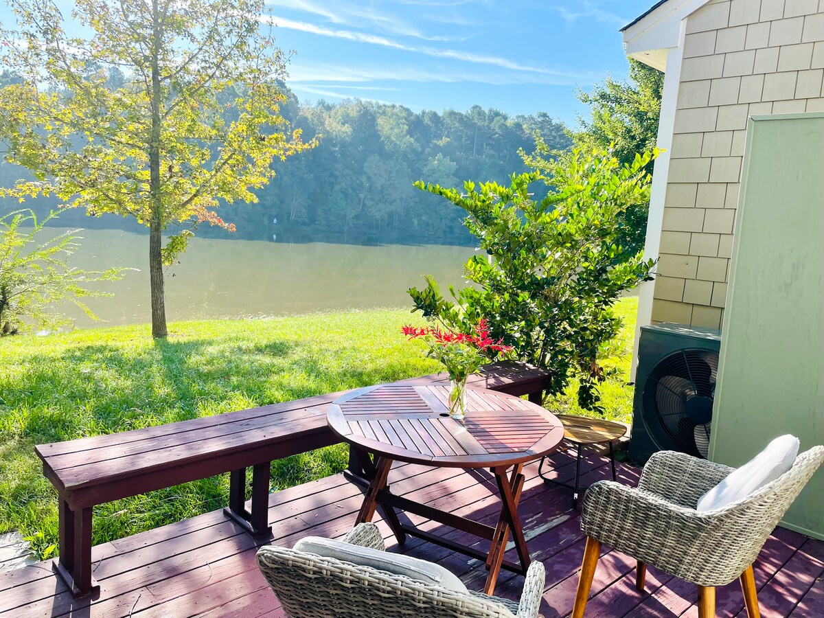 A tranquil outdoor seating area features a round wooden table surrounded by two woven chairs and a long bench. A vase with fresh flowers sits on the table, and the serene lake view is framed by lush greenery and distant trees.