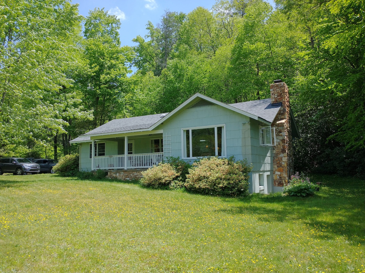 The cottage is set in a lush green landscape, surrounded by trees that provide a natural canopy. A stone chimney adds character to the exterior, while a front porch invites relaxation. The well-kept yard features patches of wildflowers and ample space for outdoor activities.