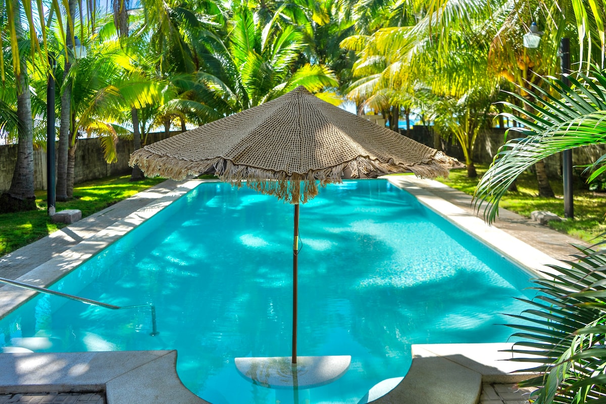 A large swimming pool is framed by lush palm trees, creating a serene outdoor space. A shaded umbrella stands at the pool's edge, inviting relaxation under the tropical sun. The clear water reflects the vibrant greenery surrounding the area.