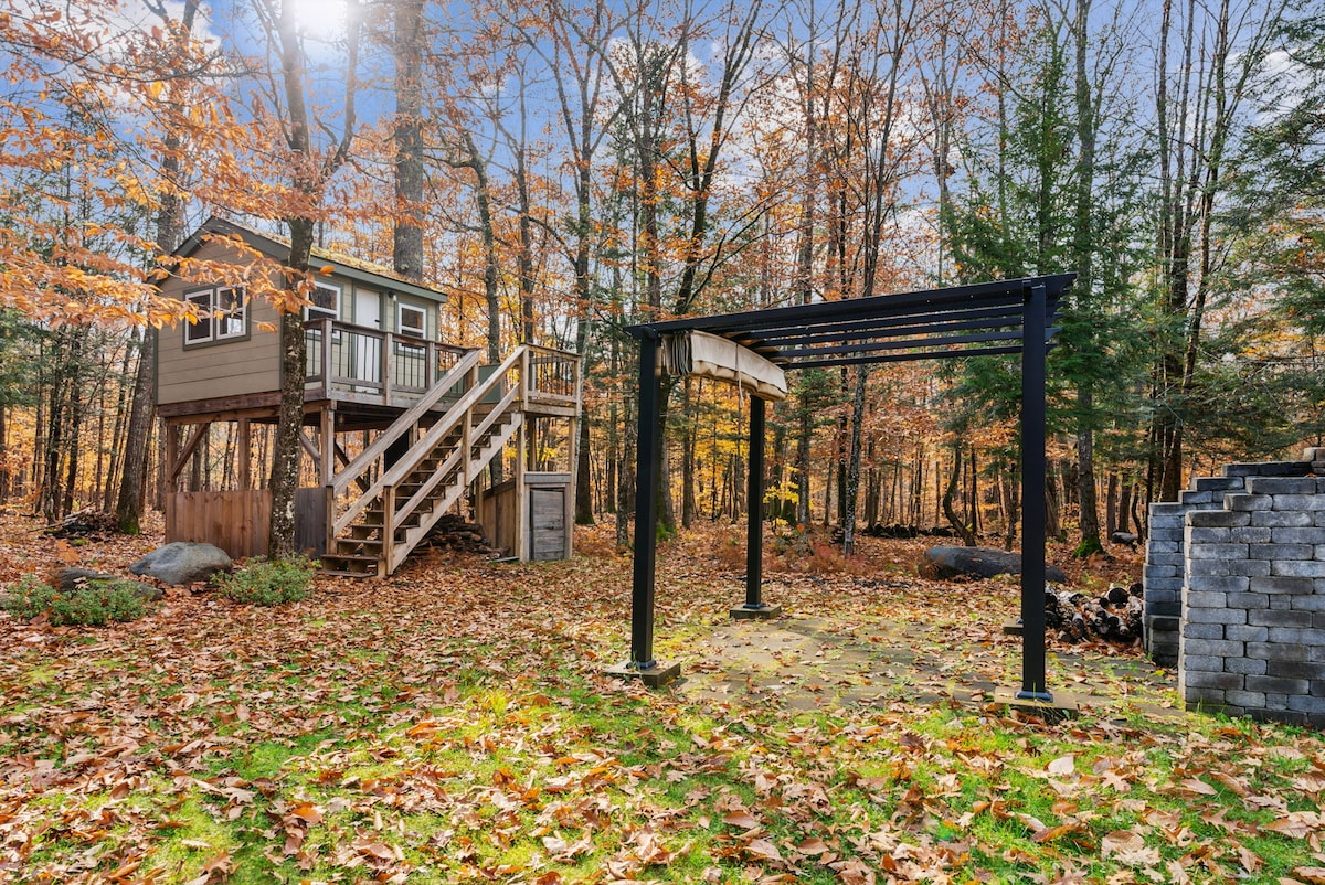 A charming treehouse nestled among trees showcases a stairway leading up to its entrance. Leaves blanket the ground, highlighting the changing seasons. A black structure stands nearby, complementing the natural setting, while stacked firewood is visible to the side, enhancing the outdoor atmosphere.