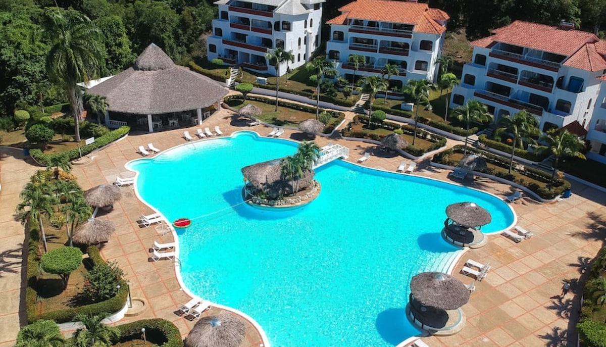 A large, inviting swimming pool is surrounded by lush greenery and well-maintained landscaping. Palm trees sway gently nearby, while shaded lounge areas with thatched roofs offer relaxation. Comfortable sun loungers are positioned around the pool, creating a serene environment ideal for relaxation.